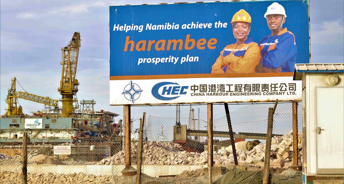 Ein großes blauweißes Schild mit der Personen drauf, die jeweils einen Bauarbeiterhelm tragen. Hinter dem Schild sieht man große Steinhaufen und hohe Kräne.