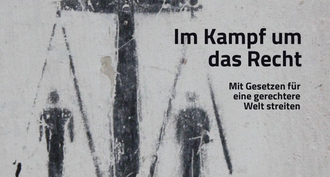 Auf einer grauen Wand ein schwarzer Print mit Justizia-Waage