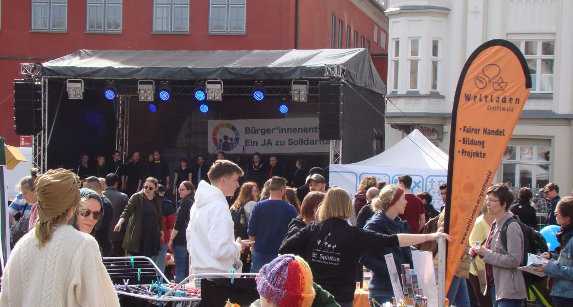 Viele Menschen auf einem Straßenfest. Im Hintergrund ein Chor auf einer Bühne.