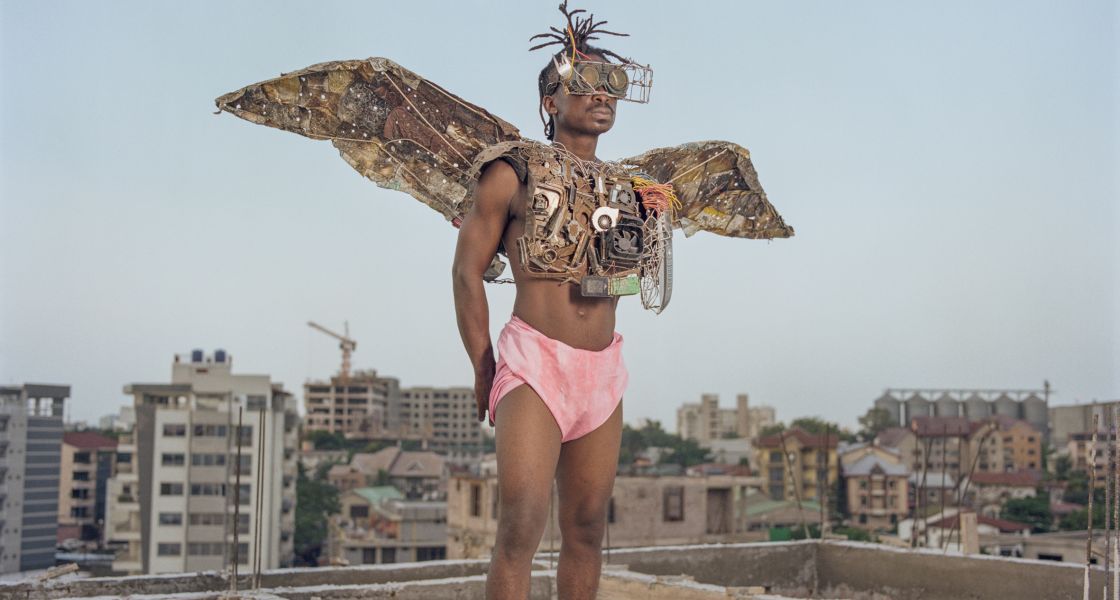 Ein schwarzer Mann steht in rosa Shorts und mit aus Schrott gebauten Flügeln auf einem Dach im Stadtviertel Kitambo