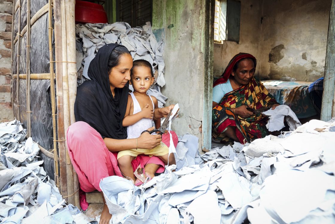 Eine Frau sitzt auf einem Szapel helles Leder. Ein Kind sitzt auf ihrem Schoß. Die Frau schneit Leder. Durch eine Wand getrennt sitzt noch eine Frau und schneidet Leder.