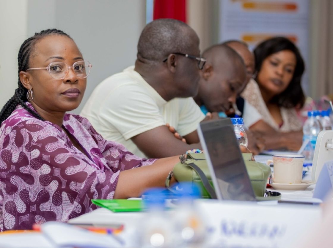 Eine Frau sitzt an einem langen Tisch und guckt in die Kamera, hinter ihr sitzen vier weitere Personen