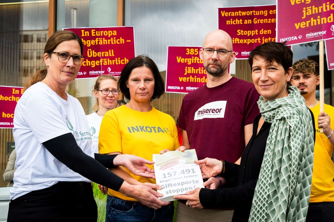Vier Menschen halten gemeinsam ein Papierpaket in den Händen. Dahinter stehen andere Menschen mit Protestschildern