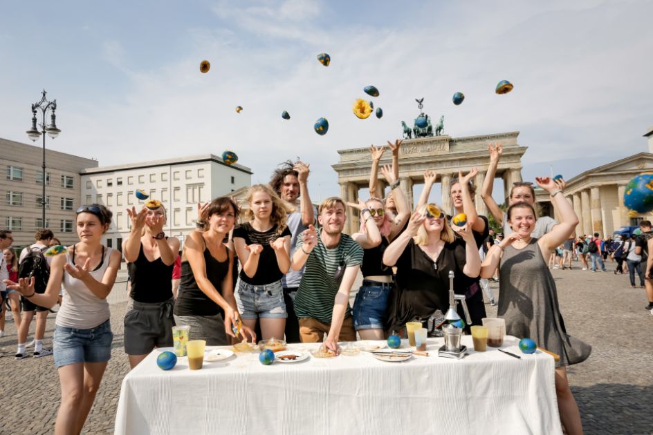 Ene Gruppe junger Menschen vor einem Tisch und im Hintergrund das Brandenburger Tor