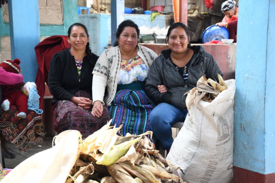 Drei sitzende Frauen blicken in die Kamera. Vor ihnen liegen Maiskolben.