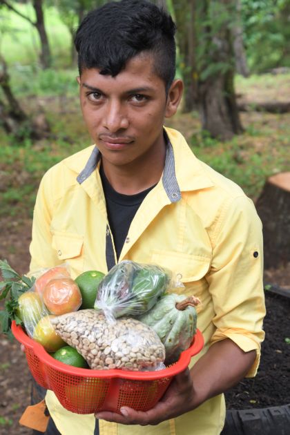 Ein junger Mann mit gelbem Hemd hält verschiedene Gemüse in den Händen und schaut direkt in die Kamera