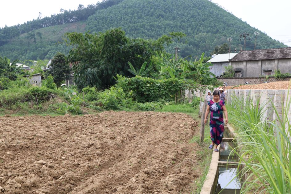 Eine Frau im Kleid balanciert auf einem Steg neben einem Acker und einem Bewässerungsgraben