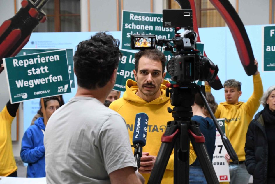 Ein Mann, der Mikrofon hält, und ein anderer Mann, de rins Mikrofon spricht. Dahinter sind Menschen mmit Protestschildern zu sehen.