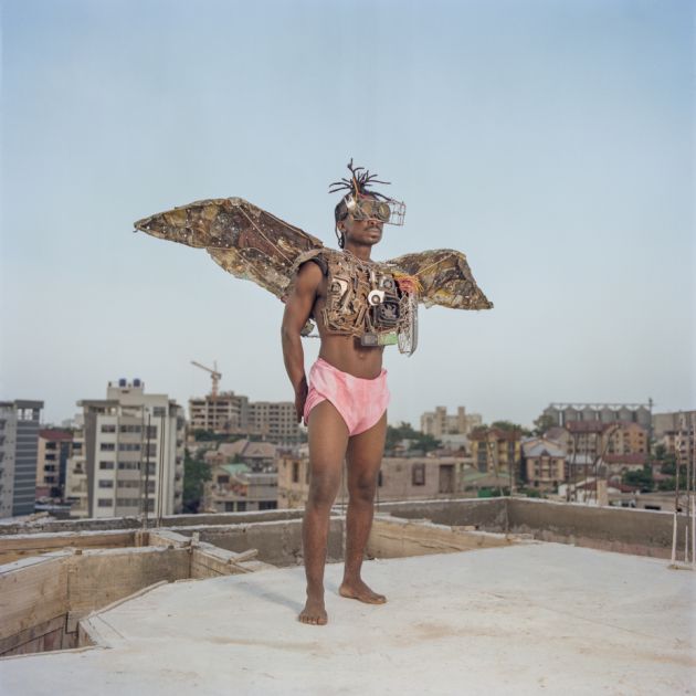 Ein schwarzer Mann steht in rosa Shorts und mit aus Schrott gebauten Flügeln auf einem Dach im Stadtviertel Kitambo