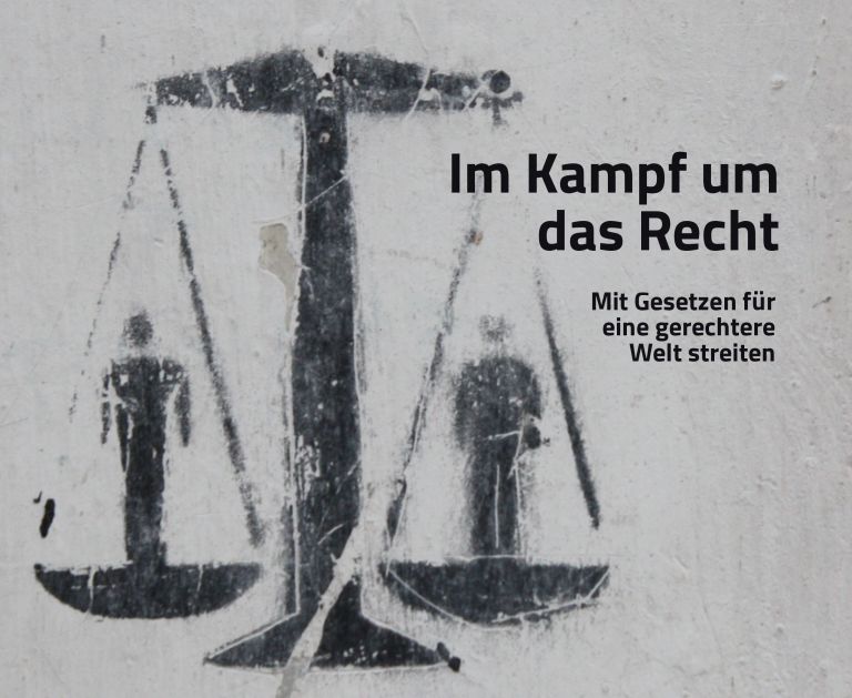 Auf einer grauen Wand ein schwarzer Print mit Justizia-Waage