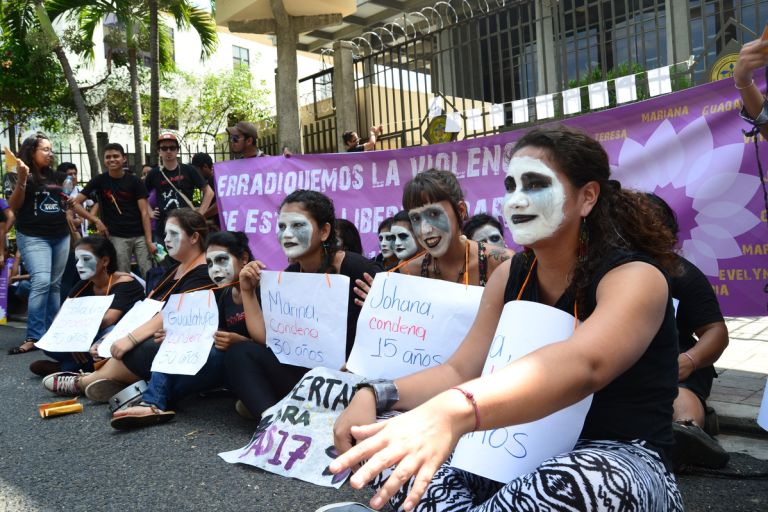 Mehrere Menschen sitzen auf dem Boden. Ihre Gesichter sind weiß geschminkt. Um den Hals haben sie Protestschilder umhängen.