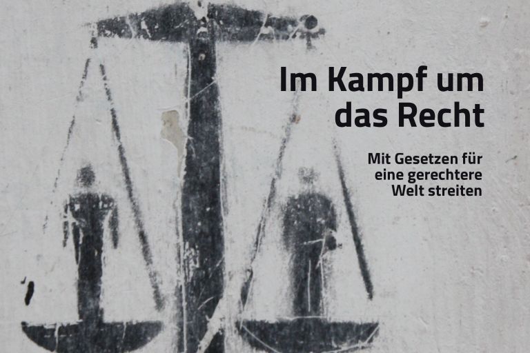 Auf einer grauen Wand ein schwarzer Print mit Justizia-Waage