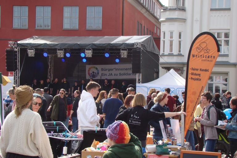 Viele Menschen auf einem Straßenfest. Im Hintergrund ein Chor auf einer Bühne.