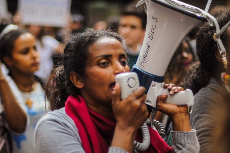 Eine Frau mit rotem Schal und einem Megafon spricht in das Gerät rein
