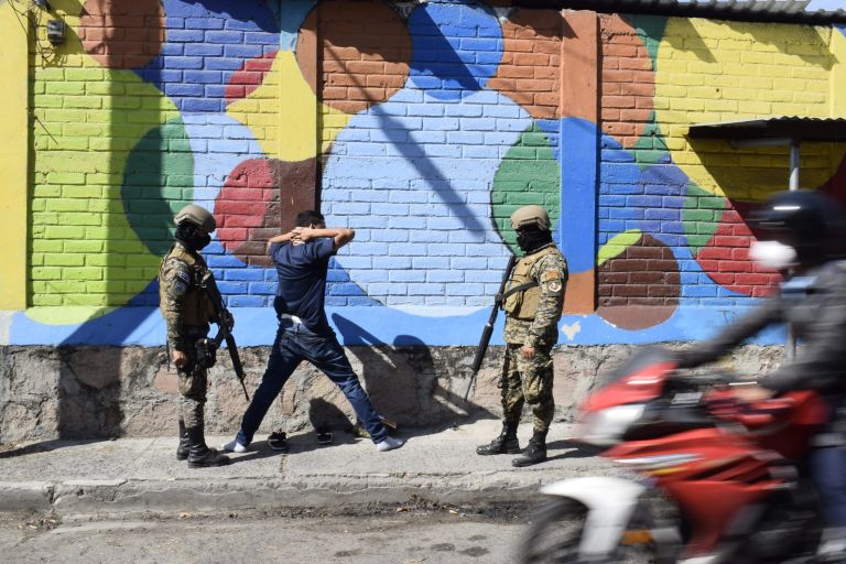 Ein Mann mit erhobenen Händen hinter dem Kopf und zwei uniformierten Militärs daneben