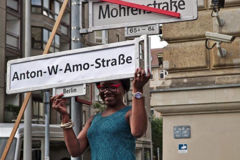 Eine Schwarze Aktivistin hält ein Straßenschild mit Namen Anton-W-Amo-Straße in die Höhe, darüber ist das Straßenschild der M*-Straße rot durchgestrichen